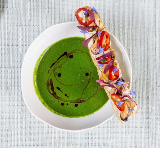 An overhead shot of a bowl of bright green soup with a dark oil drizzle. A long baguette slice sits on the edge, decorated with mussels, red chili rings, and delicate purple and blue floral garnishes. A corner of another plate with a small appetizer is visible in the top left.