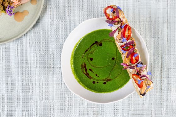 An overhead shot of a bowl of bright green soup with a dark oil drizzle. A long baguette slice sits on the edge, decorated with mussels, red chili rings, and delicate purple and blue floral garnishes. A corner of another plate with a small appetizer is visible in the top left.