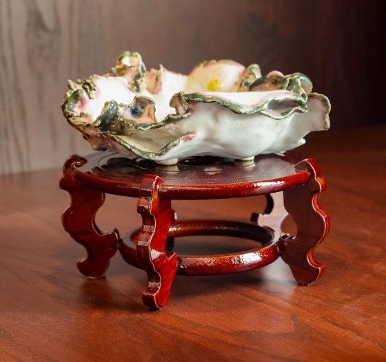 A close-up of an empty, highly ornate Kusamono pot displayed on a dark red, polished wooden stand with curved, decorative legs. The ceramic pot is designed to resemble a delicate, fluted seashell or ruffled flower blossom, featuring a white interior and a heavily textured rim highlighted with soft pink and green glazes.