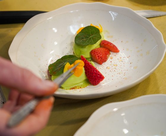 An over-the-shoulder close-up of a chef using kitchen tweezers to place a delicate orange edible flower petal onto a plated dish consisting of vibrant green sorbet, sliced fresh strawberries, and green nasturtium leaves in a wide white ceramic bowl.