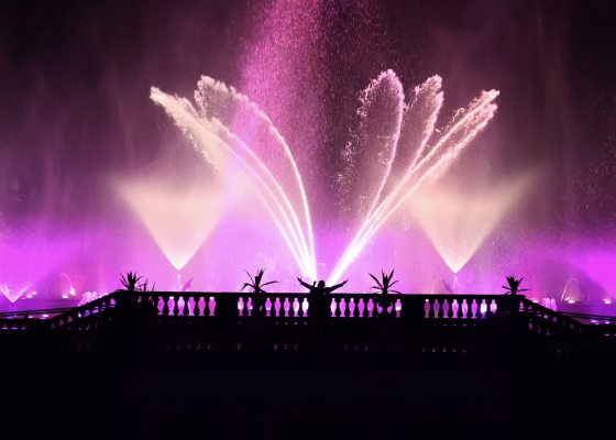 Silhouette of a person standing with arms raised on a balcony, watching a grand, fan-shaped nighttime fountain display illuminated in a deep magenta hue.