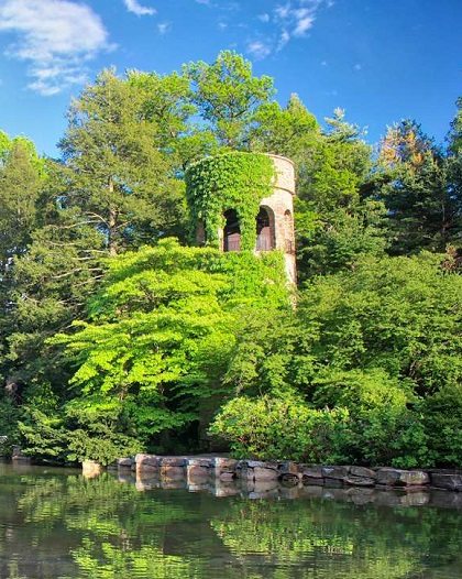 a small pond surrounded by large green trees, a waterfall, and a stone tower in the distance