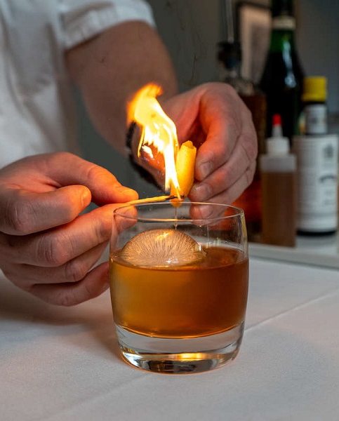 a glass of alcohol with a set of hands lighting a lemon peel on fire 