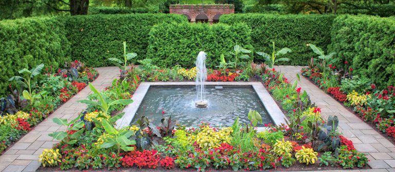 a square fountain surrounded by flower beds in bloom and enclosed by a green hedge