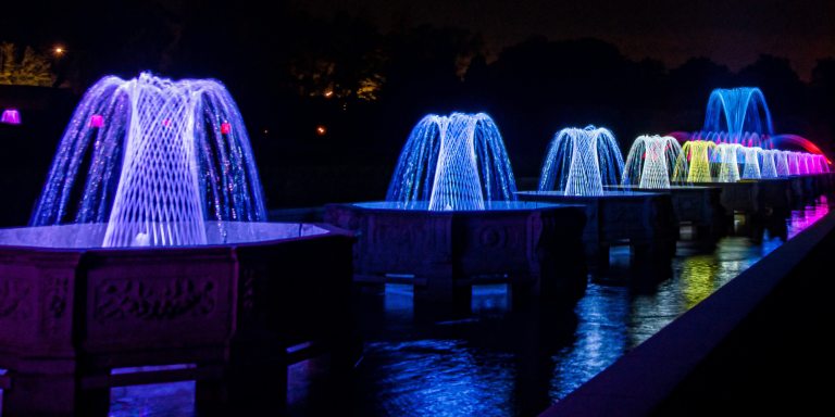 a row of basketweave fountains in octagonal basins reflect their blues, pinks, and purples in the water at night
