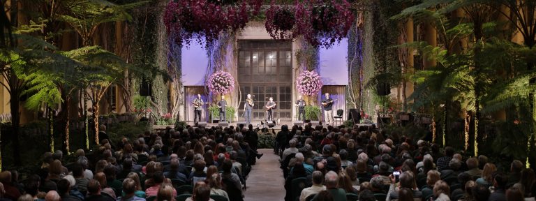 performers on a stage in an indoor garden setting, with audience in foreground