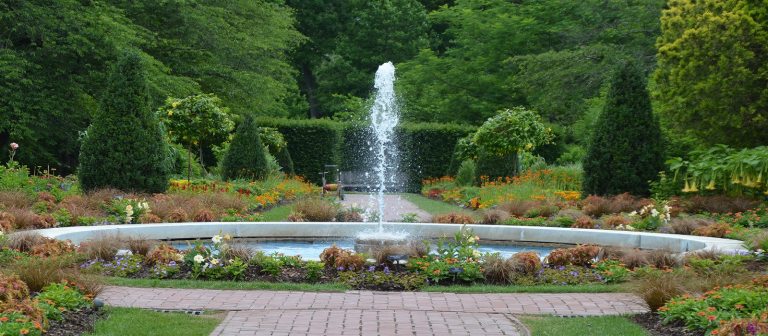 a round fountain set in the middle of two paths of gardens in bloom on either side of it