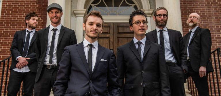 Six adults in suits standing in front of a brick building.