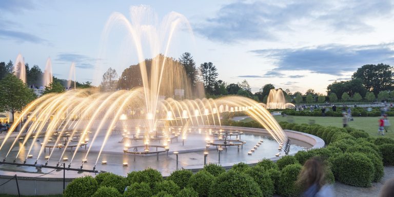 a central fountain surrounded by jets of water spraying toward it, all lit by white light