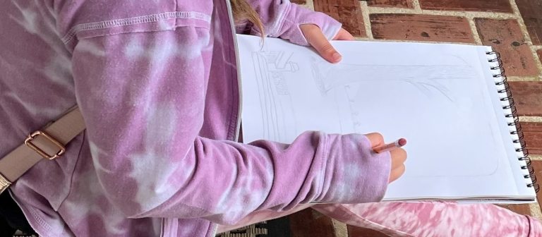 a person with pink top and pants draws with a pencil in a spiral sketchbook