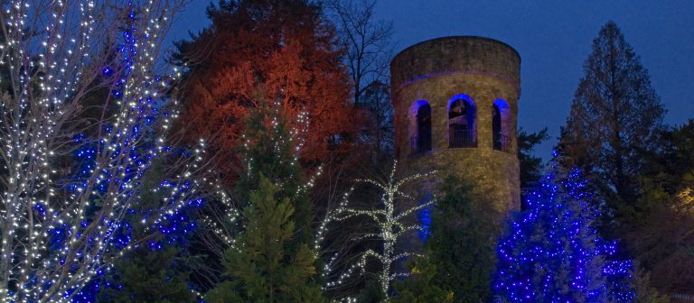 a stone tower with arched openings outlined in blue lighting against a darkening sky, with holiday-lit trees in the foreground