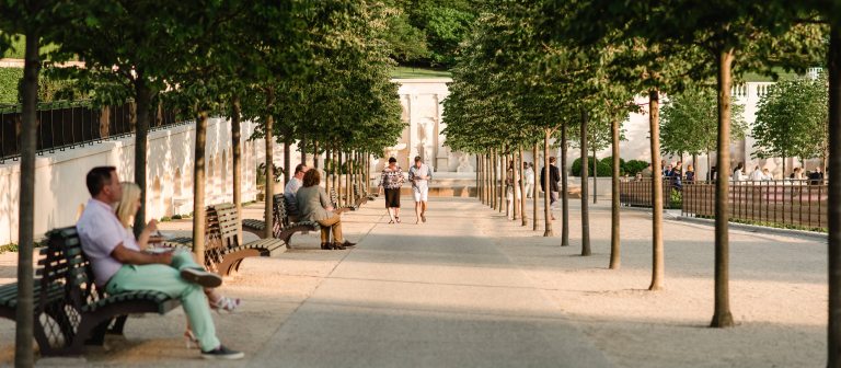 a wide view down a white walkway lined with trees and garden benches, looking toward a white stone fountain wall