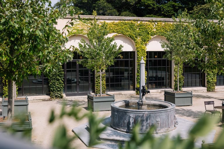 a circular fountain adorned with ornate sculptures sits in a courtyard surrounded by arching walls and green plants