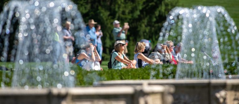 a group of people sitting on a bright green lawn behind a spray if water coming from two fountains