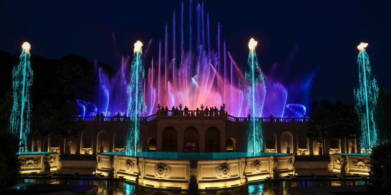 pink, purple, and blue fountains dance in the background, with 4 jets of green water topped by flames in the foreground