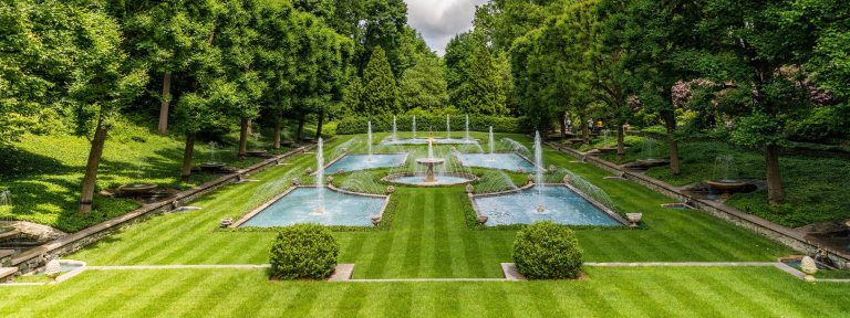 fountains rise from rectangular blue pools, surrounded by green lawn and a backdrop of trees