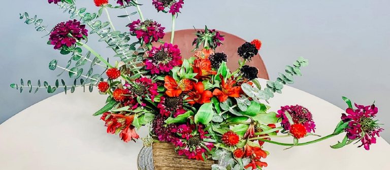 An asymetrical floral arrangement in a gold vase sitting on a white table cloth.