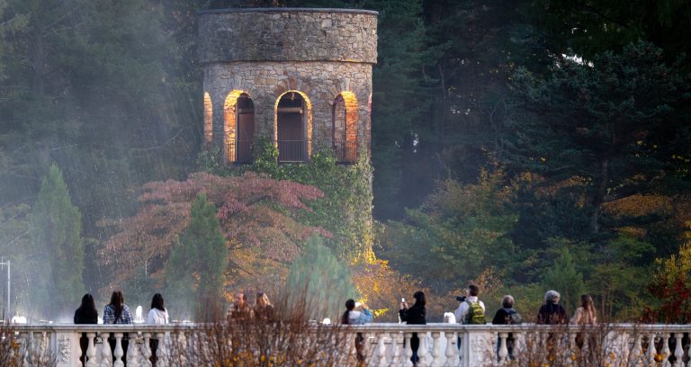 A view of the illuminated stone Chimes Tower, seen through fountain spray amid colorful fall foliage, with guests watching and some taking pictures from an overlook with stone rail and columns.