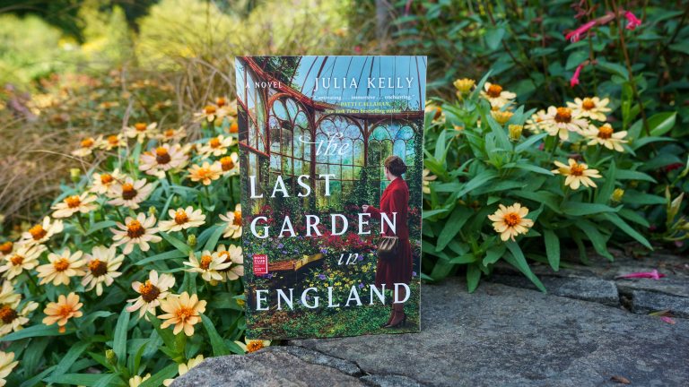 A book entitled "Last Garden in England" is propped on a stone garden wall, backed by yellow flowers, and a mix of light to dark green foliage.