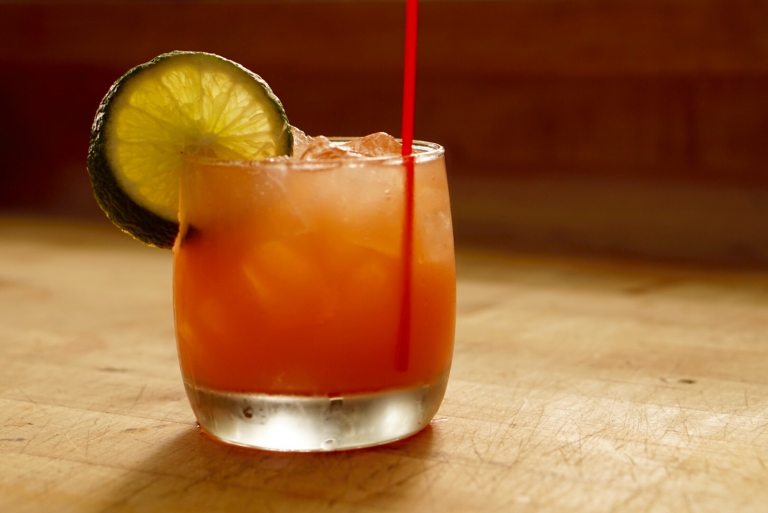 A peach colored cocktail resting on a table with lime wedge and red stirrer.