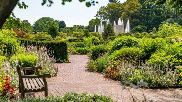 A brick walkway with a wooden bench winds through green garden beds with fountains in the distance.