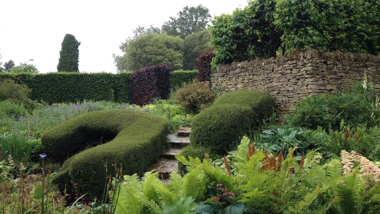 A lush garden with a stone wall, topiary, and flowery shurbs.