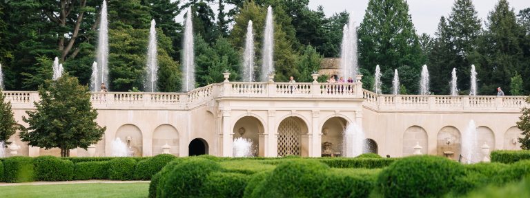 Multiple fountains rise up from two levels of a white stone facade and promenade, with large green trees and a glimpse of a stone carillon tower in the background; and smaller trees, green lawn, and boxwood in the foreground.