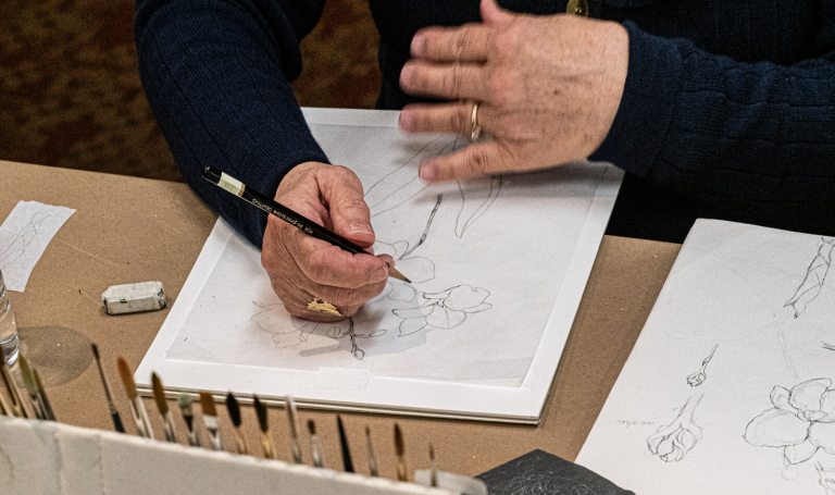 A person seated at a table using a pencil to draw a plant on white paper.