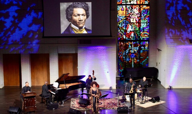 Performers sing and play instruments on stage, below a screen on which is projected an image of Frederick Douglass, next to a floor-to-ceiling stained glass window.