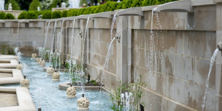 Diagonal shot of stone wall with fountains spouting water into a long blue-tiled basin below, in which the spray of water from stone floral fountain features mimics the habit of the green papyrus plants nearby.