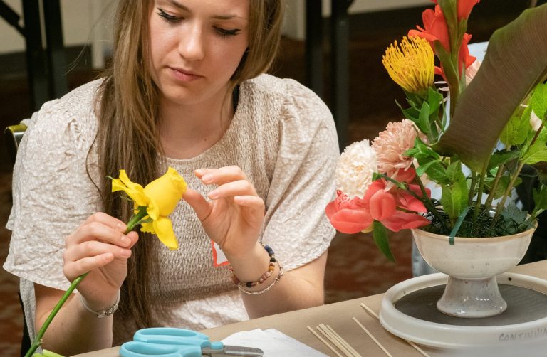 A seated person creating a floral arrangemetn, holding a yellow rose.