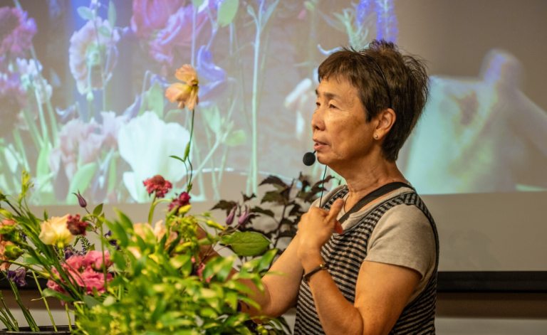 A person standing in front of a projection screen, talking into a microphone, next to bundles of cut flowers.