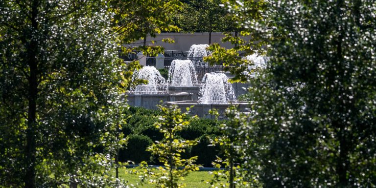 Multiple fountains in basketweave formation rise from octagonal stone basins, seen through a tapestry of green trees.