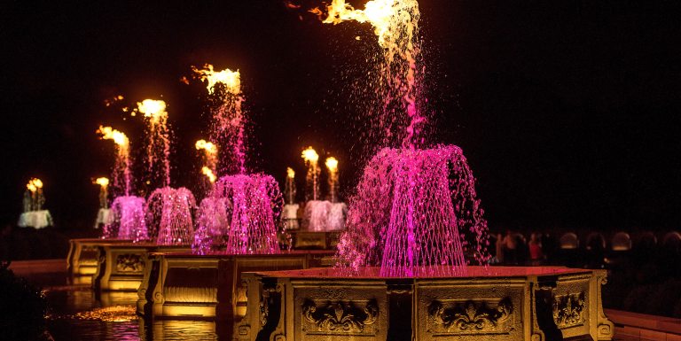 A row of octagonal stone basins at night, with fountains in basketweave formation, lit up in fuchsia and topped by fire.