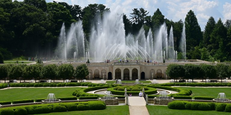 Wide view of a fountain garden with green lawn and boxwood hedges in front of a large stone facade with multiple arches, atop which jets of water reach toward the sky against a backdrop of green trees.