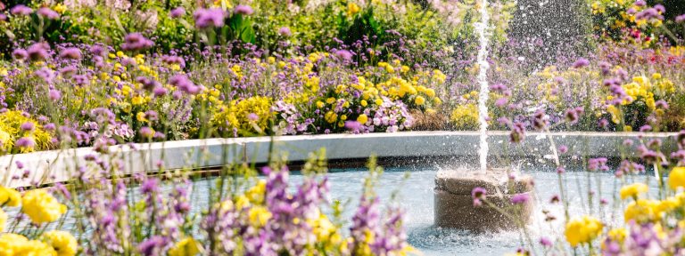 A single jet of water shoots up from a round fountain surrounded by pink and yellow flowers.