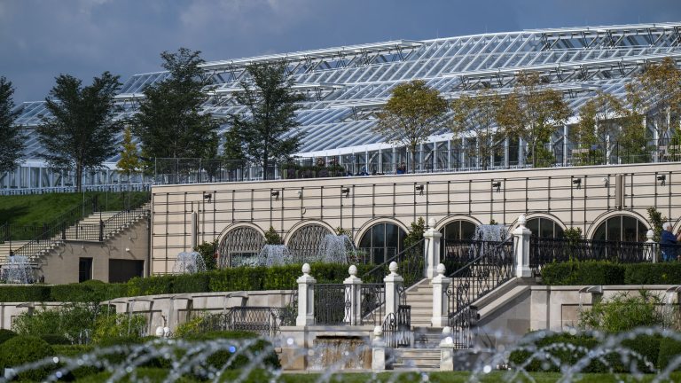 A new large conservatory complex spans three visible levels against a blue sky reflected by the greenhouse windows, with fountains in the foreground.