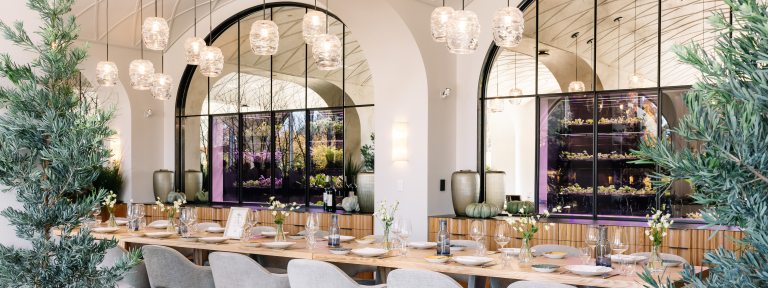 A long oak table is set with white dinner plates and clear glasses. In the background are arched mirrors and growing herbs. 