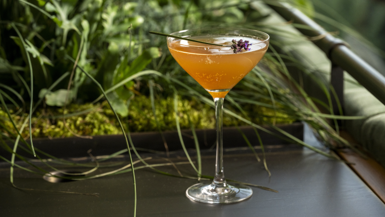 Closeup of amber colored cocktail in stemmed glass, topped with purple floral sprig.