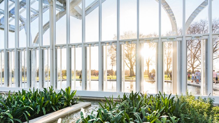 A view from the inside looking out of a glass house, with green plants and fountains in the foreground, and a view through the windows of golden hour, trees, and people approaching.