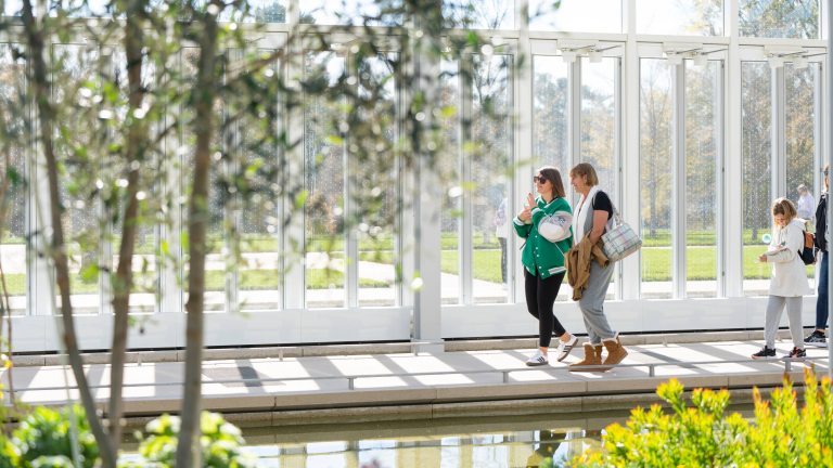 Guests stroll along a sunlit indoor path in a glass house, with water and green plants in view.