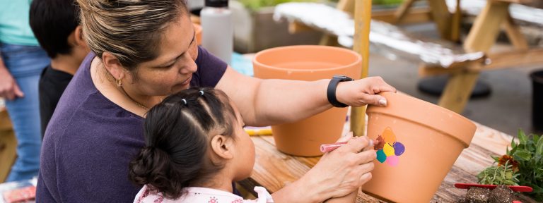 An adult and child paint a multi-colored flower on a large clay pot.