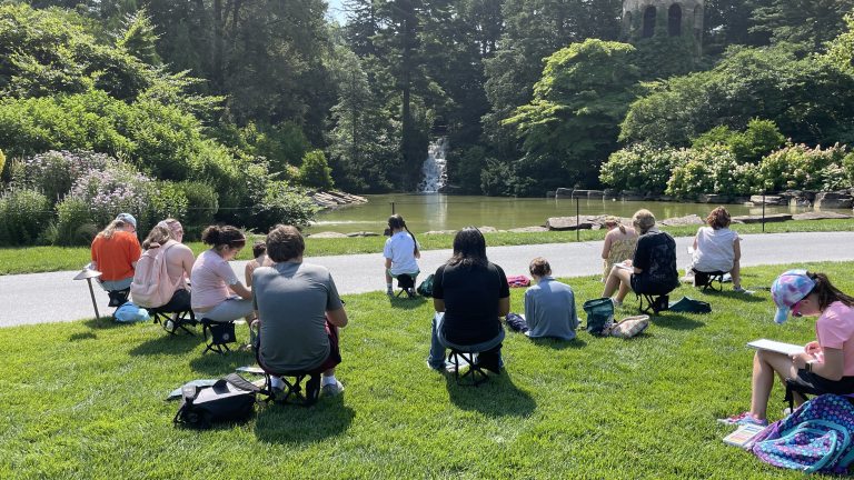 Young people, seated on folding garden chairs, sketch a landscape containing a pond, waterfall, and stone tower amid trees and gardens.