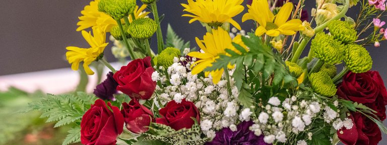 A close up of a floral arrangement featuring mini red roses, green foliate, and yellow flowers.