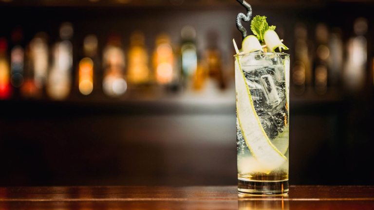 A clear cocktail, garnished with cucumber, onto of a dark wood bar. 