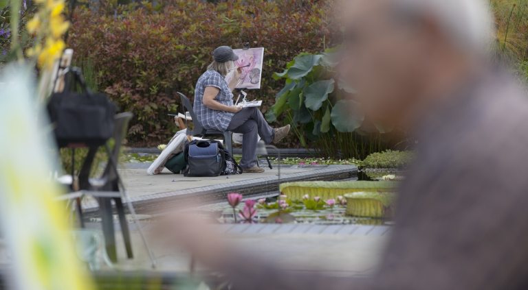 Two people, one in the foreground and blurry, and one further away, seated at an easel painting images of waterlilies at Longwood Gardens.