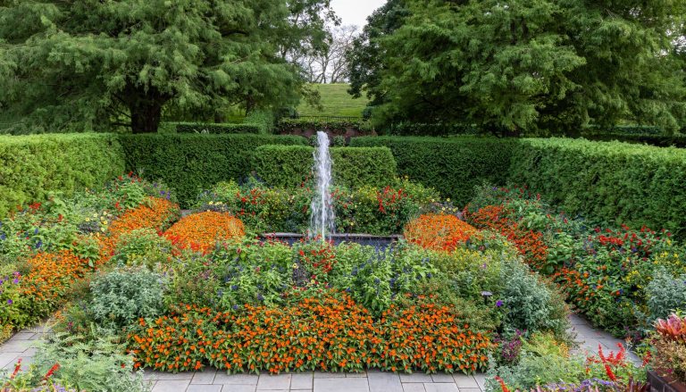 A square garden, with a square fountain in the center, and lush garden beds surrounding it, featuring orange and red blooms.