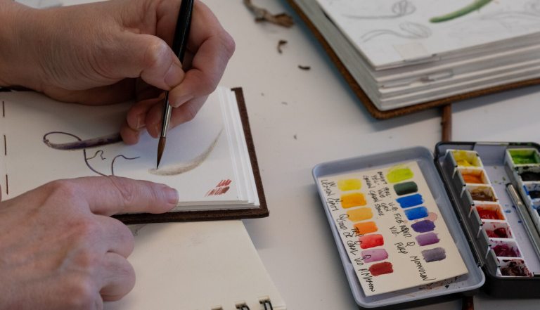 A person painting in a sketchbook with an open color palette next to them.