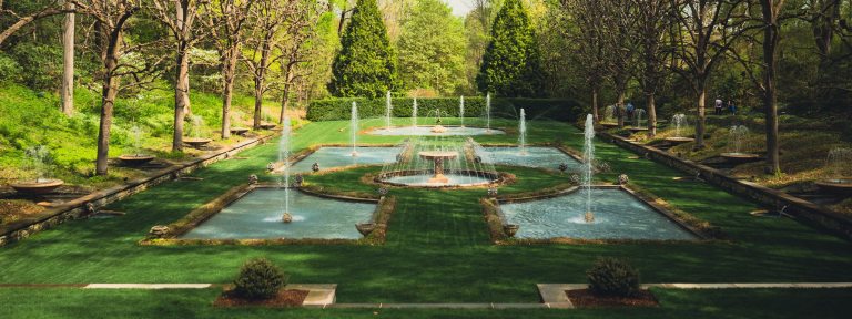 Wide view of fountain garden on a green lawn bordered by trees, in half light and half shadow.
