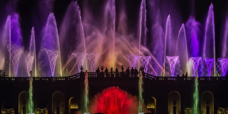 Expansive view of illuminated fountain show with large upper fountains in shades of purple, and lower fountains in green and red.